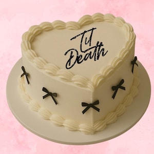 May include: A heart-shaped cake with white icing and cream swirls around the edges. The cake features black bow decorations and the words "Til Death" in black script. It rests on a white plate, set against a pink background.