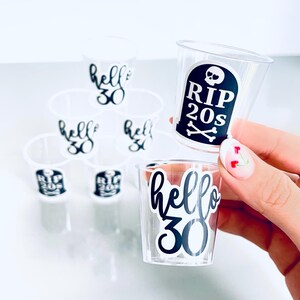 May include: Clear plastic shot glasses with black text. Some glasses say "hello 30" and others say "RIP 20s" with a skull and crossbones. The shot glasses are arranged in a pyramid shape.