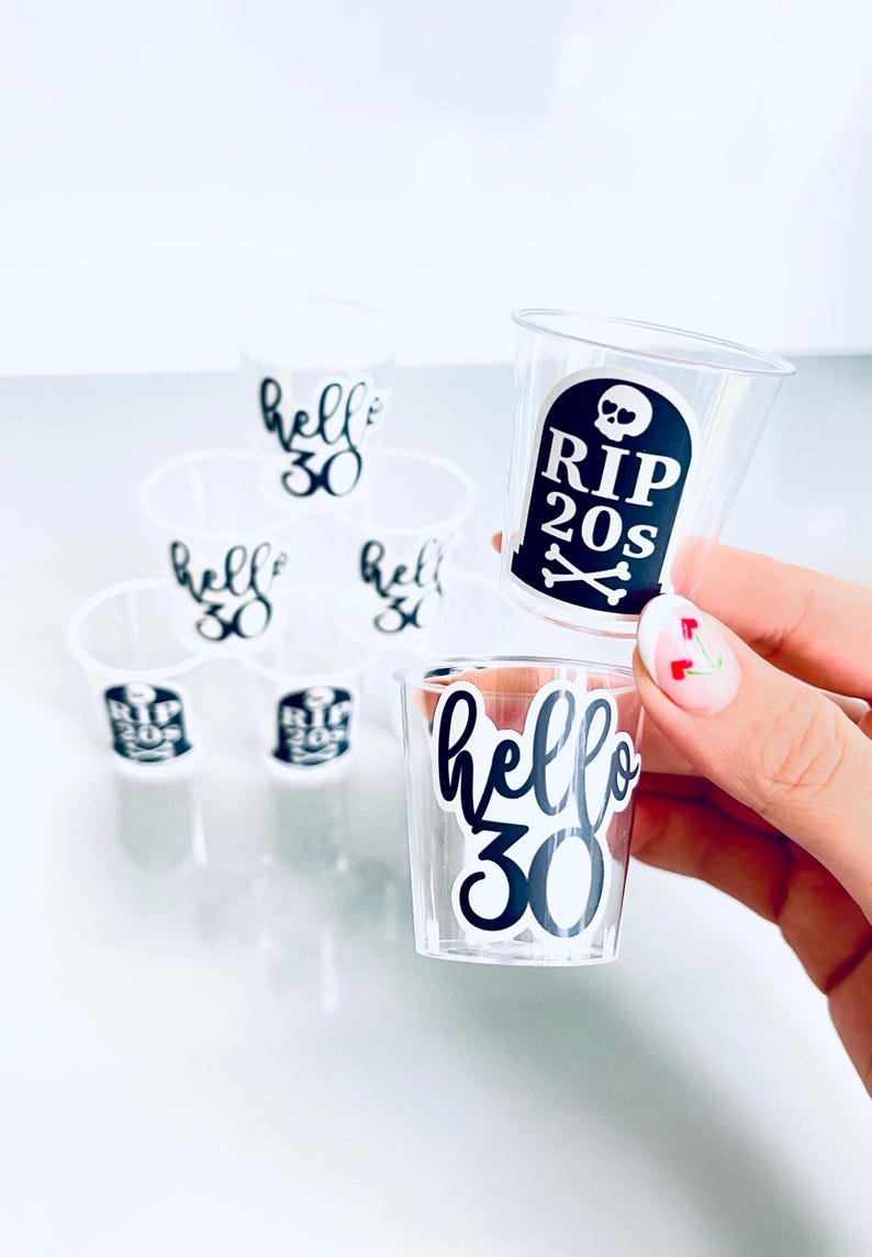 May include: Clear plastic shot glasses with black text. Some glasses say "hello 30" and others say "RIP 20s" with a skull and crossbones. The shot glasses are arranged in a pyramid shape.