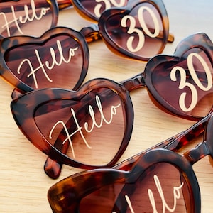 May include: Multiple pairs of heart-shaped sunglasses with a brown tortoiseshell pattern. The lenses feature white text printed on them, with some lenses displaying the word Hello in a cursive script and others displaying the number 30.