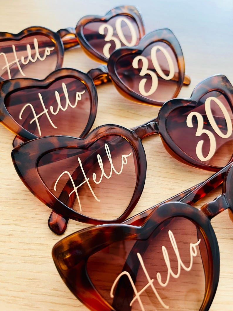 May include: Multiple pairs of heart-shaped sunglasses with a brown tortoiseshell pattern. The lenses feature white text printed on them, with some lenses displaying the word Hello in a cursive script and others displaying the number 30.