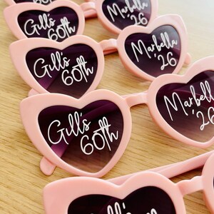 Pode incluir: Uma cole&ccedil;&atilde;o de &oacute;culos de sol em forma de cora&ccedil;&atilde;o cor-de-rosa claro dispostos em filas. As lentes esquerdas apresentam o texto branco Gill's 60th, enquanto as lentes direitas exibem o texto branco Marbella '26. As arma&ccedil;&otilde;es s&atilde;o de pl&aacute;stico.