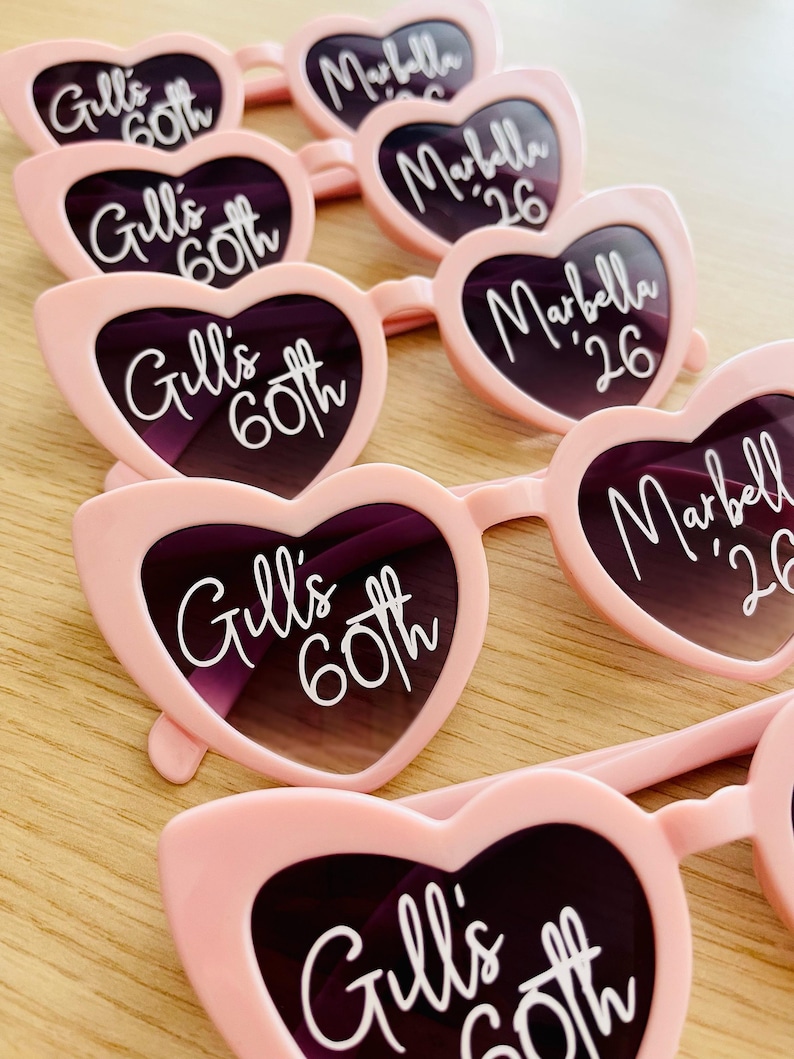 Pode incluir: Uma cole&ccedil;&atilde;o de &oacute;culos de sol em forma de cora&ccedil;&atilde;o cor-de-rosa claro dispostos em filas. As lentes esquerdas apresentam o texto branco Gill's 60th, enquanto as lentes direitas exibem o texto branco Marbella '26. As arma&ccedil;&otilde;es s&atilde;o de pl&aacute;stico.