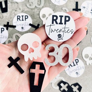 May include: A collection of black and silver glitter confetti featuring a skull, cross, coffin, and the words "RIP Twenties" and "30".