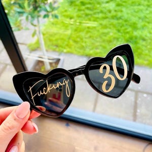 May include: Black heart-shaped sunglasses with the word "Fucking" in gold script on the left lens and the number "30" in gold on the right lens. The sunglasses are held up against a background of green foliage.