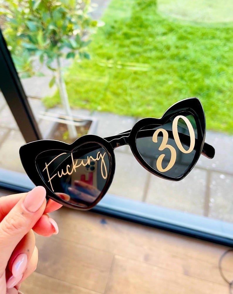 May include: Black heart-shaped sunglasses with the word "Fucking" in gold script on the left lens and the number "30" in gold on the right lens. The sunglasses are held up against a background of green foliage.