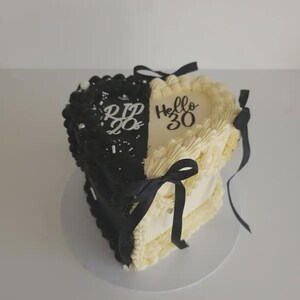 May include: A two-tiered heart-shaped cake, half black and half cream-colored, decorated with frosting. The black side says "RIP 20s" and the cream side says "Hello 30". Black ribbons adorn the cake.