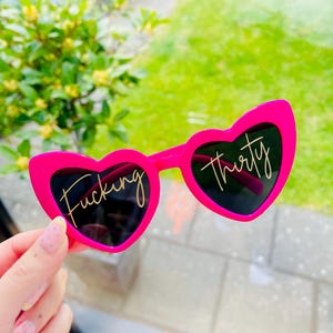 May include: Bright pink heart-shaped sunglasses with black lenses. The left lens has the word "Fucking" in gold script, and the right lens has "thirty". The sunglasses are held up against a background of green grass and foliage.