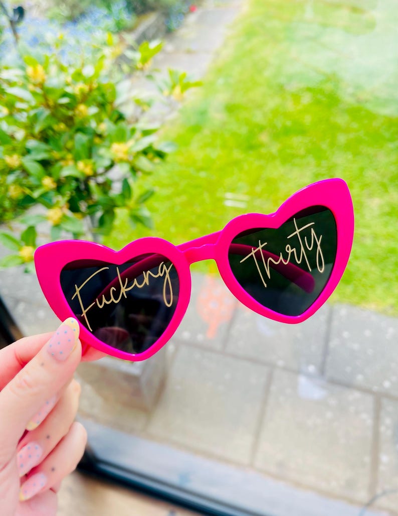 May include: Bright pink heart-shaped sunglasses with black lenses. The left lens has the word "Fucking" in gold script, and the right lens has "thirty". The sunglasses are held up against a background of green grass and foliage.