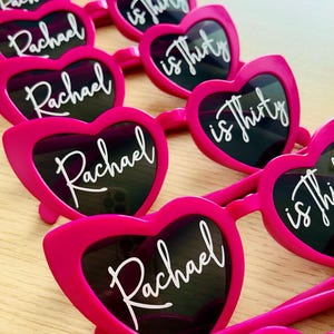 Pode incluir: Uma cole&ccedil;&atilde;o de &oacute;culos de sol em forma de cora&ccedil;&atilde;o cor-de-rosa brilhante dispostos em filas. Cada par de lentes apresenta texto branco em cursivo, com o nome Rachael na lente esquerda e a frase is thirty na lente direita.