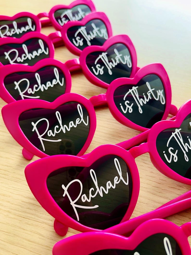 Pode incluir: Uma cole&ccedil;&atilde;o de &oacute;culos de sol em forma de cora&ccedil;&atilde;o cor-de-rosa brilhante dispostos em filas. Cada par de lentes apresenta texto branco em cursivo, com o nome Rachael na lente esquerda e a frase is thirty na lente direita.