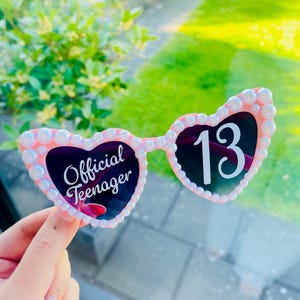 May include: Pink heart-shaped sunglasses with white pearl-like embellishments around the frames. The left lens has the text "Official Teenager" in white script, and the right lens displays the number "13" in white.