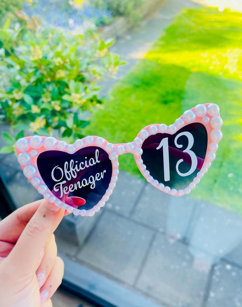 May include: Pink heart-shaped sunglasses with white pearl-like embellishments around the frames. The left lens has the text "Official Teenager" in white script, and the right lens displays the number "13" in white.