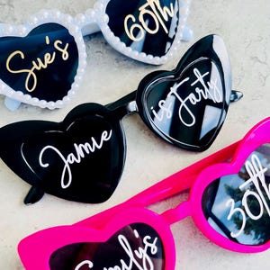 Pode incluir: Tr&ecirc;s pares de &oacute;culos de sol em forma de cora&ccedil;&atilde;o. O par superior &eacute; branco com p&eacute;rolas e texto dourado Sue's 60th. O par do meio &eacute; preto com texto branco Jamie is forty. O par inferior &eacute; rosa choque com texto branco Emily's 30th.