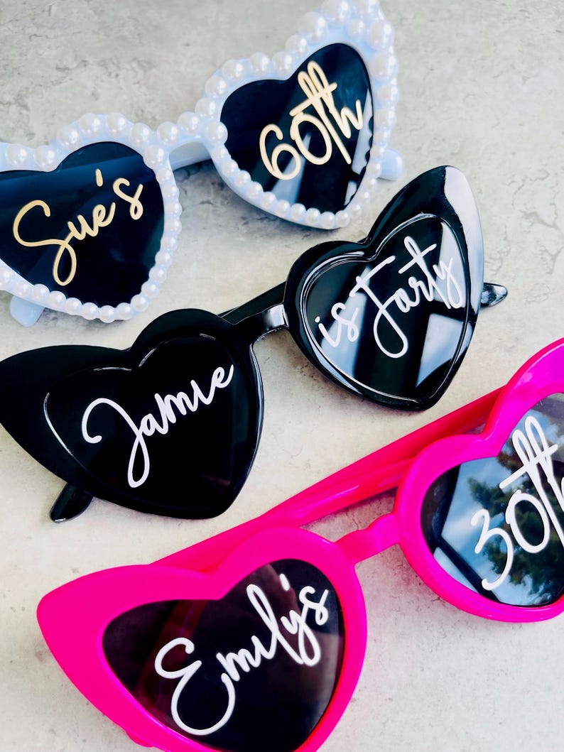 Pode incluir: Tr&ecirc;s pares de &oacute;culos de sol em forma de cora&ccedil;&atilde;o. O par superior &eacute; branco com p&eacute;rolas e texto dourado Sue's 60th. O par do meio &eacute; preto com texto branco Jamie is forty. O par inferior &eacute; rosa choque com texto branco Emily's 30th.