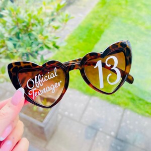 May include: Heart-shaped sunglasses with a tortoiseshell pattern. The left lens reads "Official Teenager" in white script, and the right lens displays the number "13" in white. The lenses have a gradient tint.