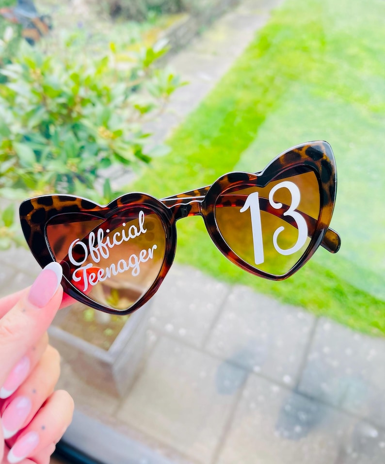 May include: Heart-shaped sunglasses with a tortoiseshell pattern. The left lens reads "Official Teenager" in white script, and the right lens displays the number "13" in white. The lenses have a gradient tint.