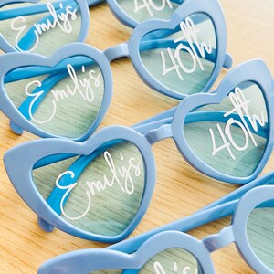 Pode incluir: Uma cole&ccedil;&atilde;o de &oacute;culos de sol azul-claros em forma de cora&ccedil;&atilde;o com texto branco impresso nas lentes. As lentes esquerdas exibem o nome Emily's em letra cursiva, enquanto as lentes direitas apresentam o texto 40th em fonte clara e negrito.