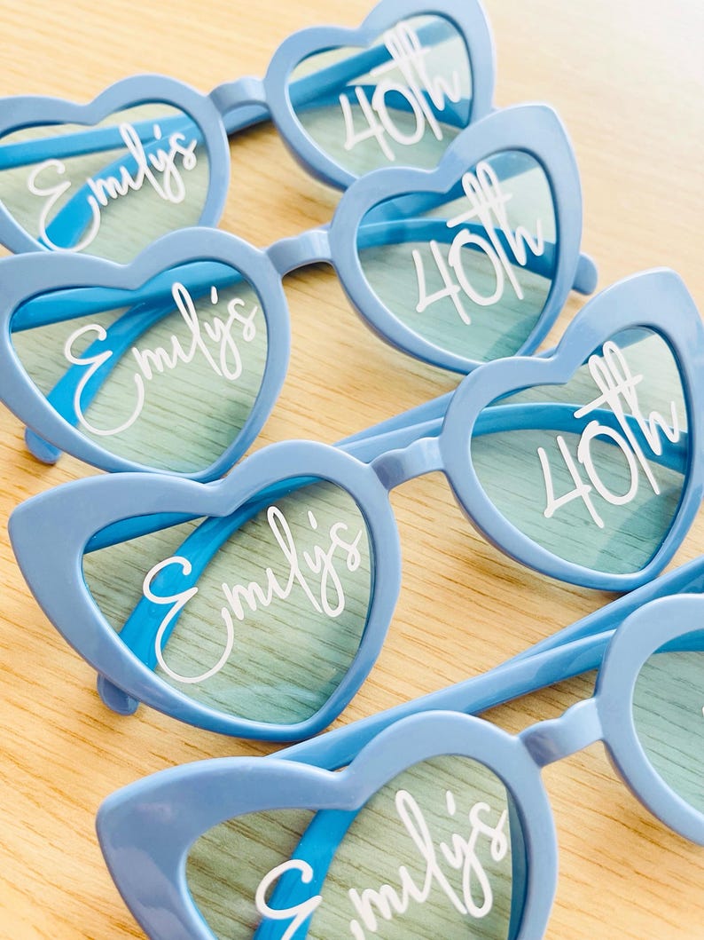 Pode incluir: Uma cole&ccedil;&atilde;o de &oacute;culos de sol azul-claros em forma de cora&ccedil;&atilde;o com texto branco impresso nas lentes. As lentes esquerdas exibem o nome Emily's em letra cursiva, enquanto as lentes direitas apresentam o texto 40th em fonte clara e negrito.