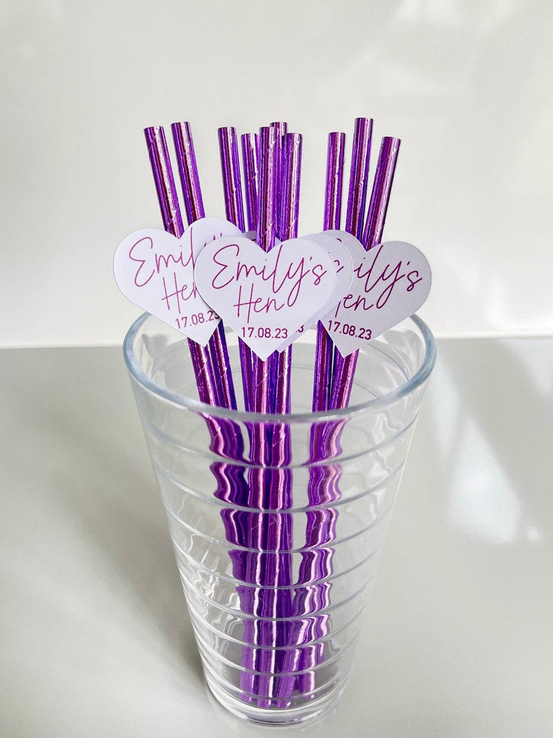 May include: Purple striped paper straws with white heart-shaped tags that say "Emily's Hen 17.08.23". The straws are in a clear glass.