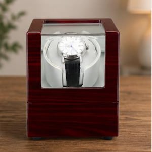 Könnte beinhalten: Ein kirschroter Uhrenbeweger aus Holz mit einer transparenten Glasplatte. Im Inneren liegt eine silberfarbene Uhr mit schwarzem Lederarmband auf einem weißen Kissen. Der Aufzieher ist für Automatikuhren konzipiert.