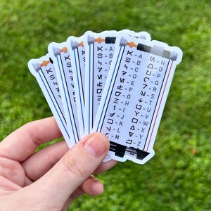 May include: A hand holds a stack of rectangular stickers. Each sticker features a white background with black, blue, and red lines. The stickers have a design with a series of symbols and letters, resembling a coded message or a sci-fi theme.