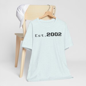 Puede incluir: Una camiseta azul claro con el texto "Est. 2002" en negro. La camiseta está colgada en una percha de madera, con una gorra de béisbol blanca apoyada en una silla de madera.