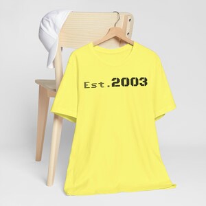 Könnte beinhalten: Ein leuchtend gelbes T-Shirt mit dem schwarzen Aufdruck "Est. 2003". Das T-Shirt hängt an einem Holzkleiderbügel. Eine weiße Baseballkappe liegt auf der Rückseite eines Holzstuhls.