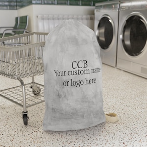 May include: A light gray laundry bag with a drawstring closure. The bag features the text "CCB Your custom name or logo here" in black. A metal laundry cart and washing machines are in the background. The bag is sitting on a speckled floor.