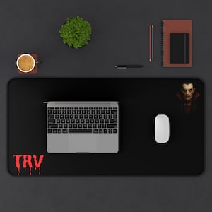 Puede incluir: Alfombrilla de escritorio rectangular negra con un portátil, un ratón y un retrato de un vampiro. La alfombrilla también presenta el texto rojo "TRV" en una fuente goteante. Una taza de café, una planta y artículos de papelería también están en el escritorio.