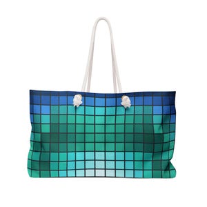 May include: Large tote bag with a pixelated design in shades of blue and green. The bag has white rope handles and white knots at the top corners. The bag is designed for carrying items.