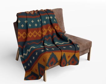 May include: A colorful knitted throw blanket draped over a brown chair. The blanket features a geometric pattern with teal, rust, gold, and cream colors. The design includes diamonds, crosses, and wavy lines, creating a cozy and decorative textile.