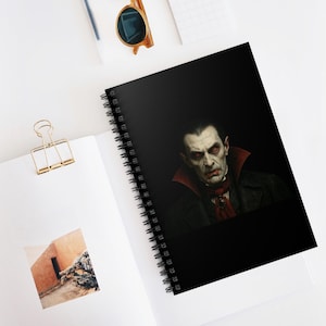 Könnte beinhalten: Spiralgebundenes Notizbuch mit einem Vampirporträt auf schwarzem Hintergrund. Der Vampir hat blasse Haut, rote Augen und einen roten Kragen. Das Notizbuch liegt auf einer weißen Oberfläche mit einer goldenen Büroklammer und Sonnenbrille.