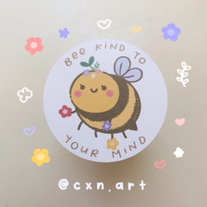 May include: A circular sticker featuring a cartoon bumblebee with pink cheeks, wearing a small floral crown, holding red and purple flowers. The text "BEE KIND TO YOUR MIND" is printed in brown around the bee. The background is white with small colorful hand-drawn flowers and hearts.