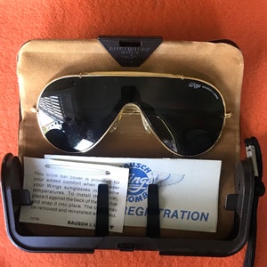 May include: A black and gold aviator style sunglasses case with a pair of black sunglasses inside. The case has a brown felt lining and a black strap. The sunglasses have a gold frame and black lenses. The case has a label that says "Wings Bausch & Lomb" and "Registration".