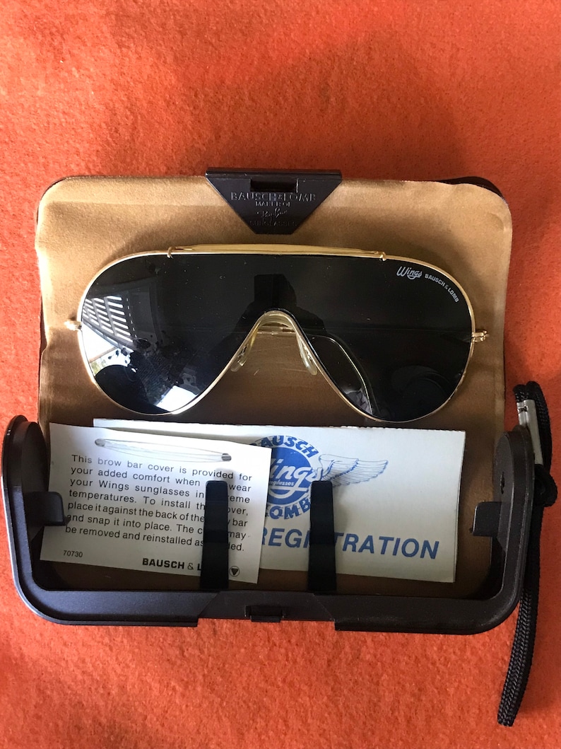 May include: A black and gold aviator style sunglasses case with a pair of black sunglasses inside. The case has a brown felt lining and a black strap. The sunglasses have a gold frame and black lenses. The case has a label that says "Wings Bausch & Lomb" and "Registration".