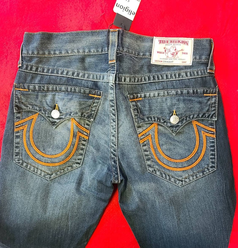 Puede incluir: Vaqueros de mezclilla azul con costuras naranjas y dise&ntilde;o de bolsillo de herradura. Los vaqueros tienen una etiqueta True Religion y cierre de bot&oacute;n. Los vaqueros son de corte recto.