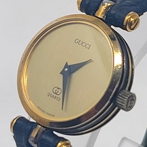 May include: A gold-toned Gucci quartz watch with a cream-colored face and black hands. The watch features a navy blue leather band and the Gucci logo. The watch face also has the word "QUARTZ" and "SWISS MADE" printed on it.