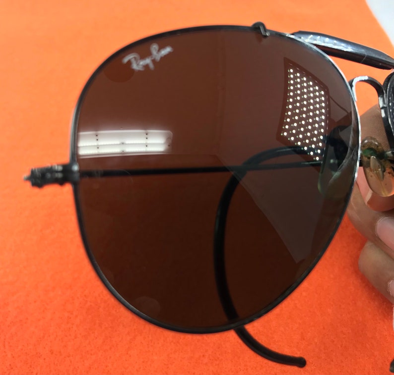 Puede incluir: Gafas de sol aviador Ray-Ban negras con lentes marrones. Las gafas se reflejan en los lentes.