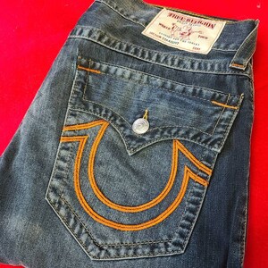 Puede incluir: Vaqueros de mezclilla azul con costuras naranjas y un gran dise&ntilde;o de herradura naranja en el bolsillo trasero. Los vaqueros tienen cierre de bot&oacute;n y una etiqueta True Religion.