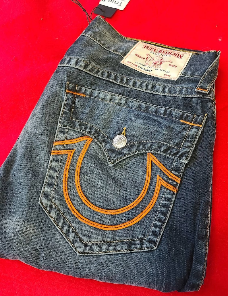 Puede incluir: Vaqueros de mezclilla azul con costuras naranjas y un gran dise&ntilde;o de herradura naranja en el bolsillo trasero. Los vaqueros tienen cierre de bot&oacute;n y una etiqueta True Religion.