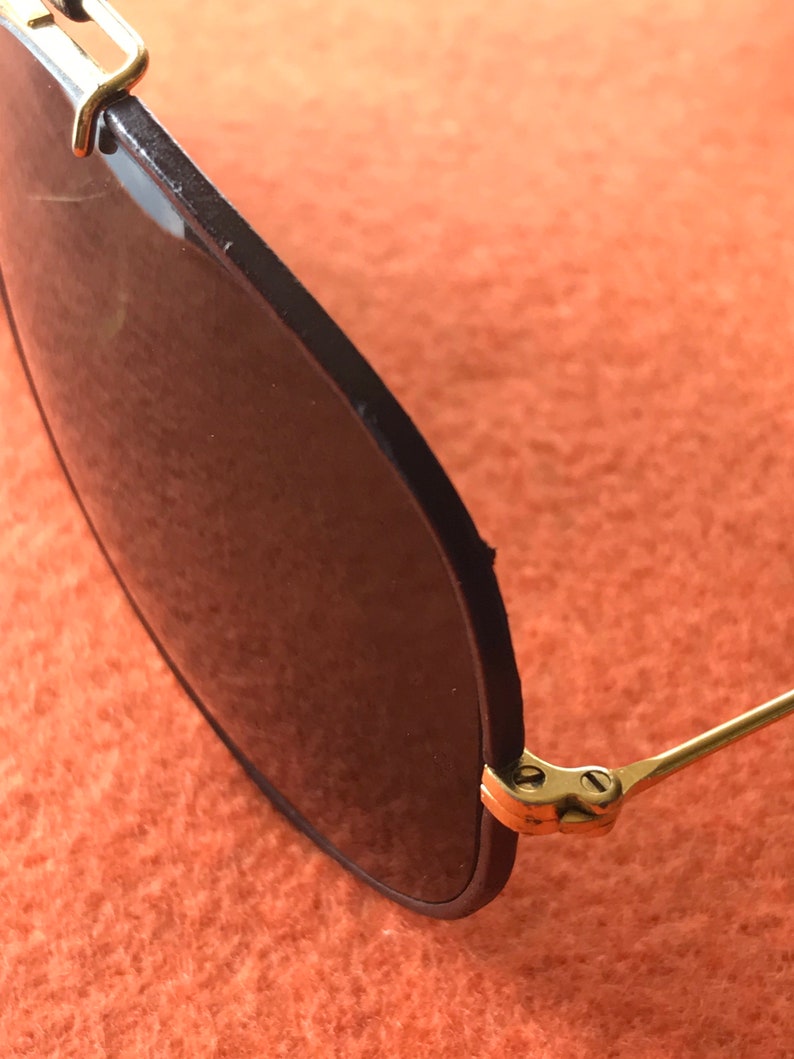Op de afbeelding: Close-up van het scharnier van een paar gouden aviator-zonnebrillen met zwarte monturen en donkere glazen.