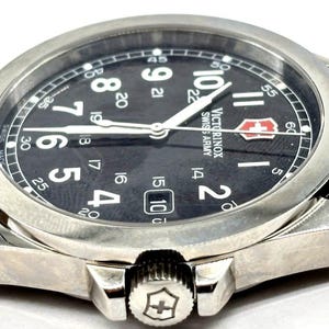 May include: Close-up of a silver-tone Victorinox Swiss Army watch with a black face. The watch features white numerals, a date window, and white hands. The watch has a stainless steel bezel and a black leather strap.