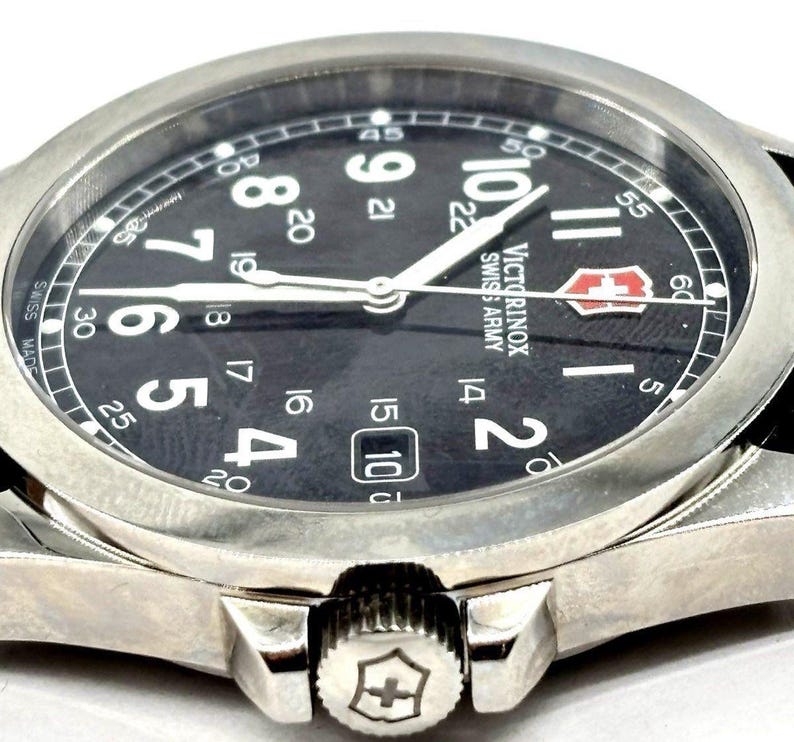 May include: Close-up of a silver-tone Victorinox Swiss Army watch with a black face. The watch features white numerals, a date window, and white hands. The watch has a stainless steel bezel and a black leather strap.