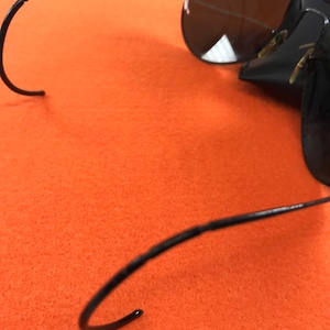 Puede incluir: Gafas de sol aviador negras con montura de metal y lentes oscuras. Las gafas est&aacute;n plegadas y las patillas est&aacute;n dobladas en forma de gancho.