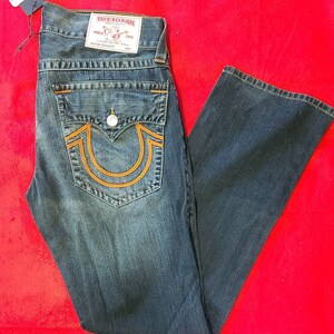 Puede incluir: Vaqueros de mezclilla azul oscuro con costuras naranjas y un dise&ntilde;o de herradura en el bolsillo trasero. Los vaqueros tienen cierre de bot&oacute;n y una etiqueta True Religion. Los vaqueros se muestran sobre una superficie roja.