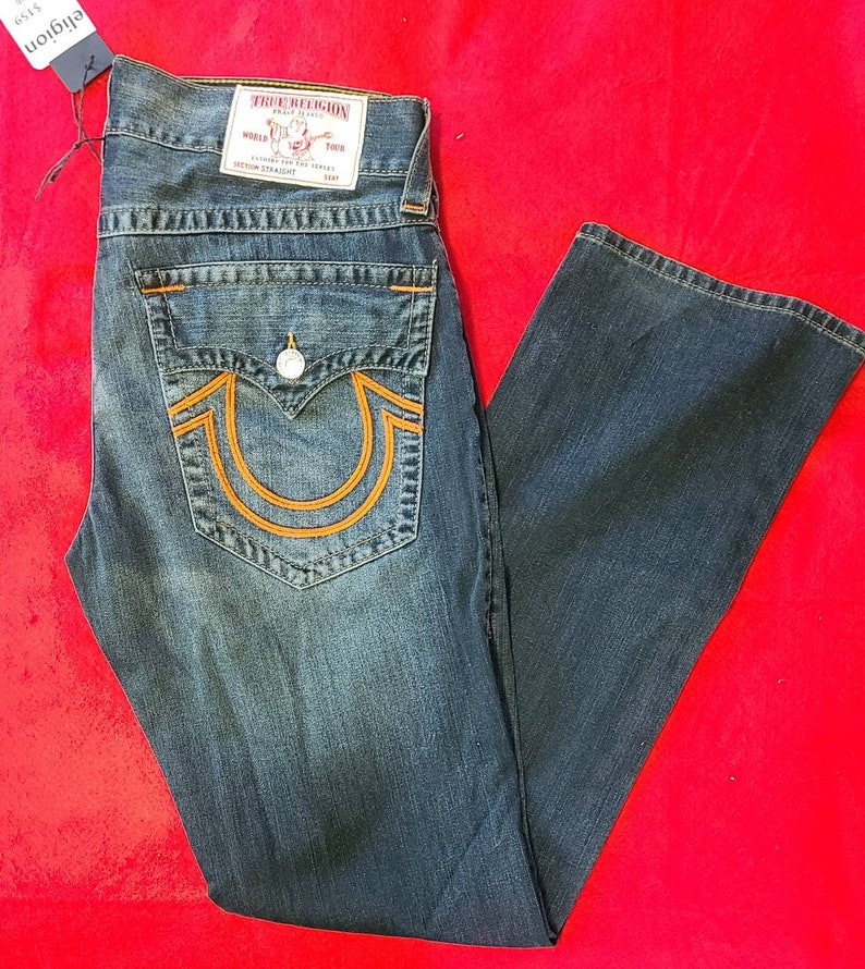 Puede incluir: Vaqueros de mezclilla azul oscuro con costuras naranjas y un dise&ntilde;o de herradura en el bolsillo trasero. Los vaqueros tienen cierre de bot&oacute;n y una etiqueta True Religion. Los vaqueros se muestran sobre una superficie roja.