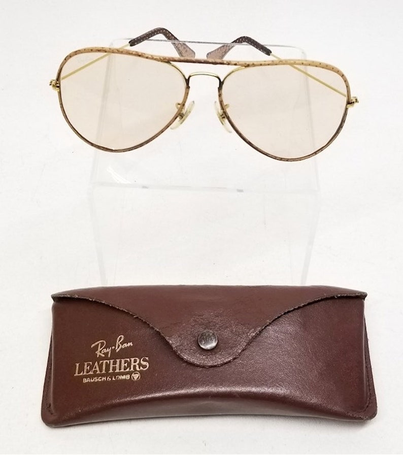 May include: Gold Ray-Ban Aviator sunglasses with a brown leather case. The case has the text "Ray-Ban LEATHERS BAUSCH & LOMB" on it.