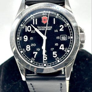 May include: A black-faced Victorinox Swiss Army watch with a stainless steel case and a black leather strap. The watch features white numerals, a date display, and the Victorinox logo. The watch is a classic design.