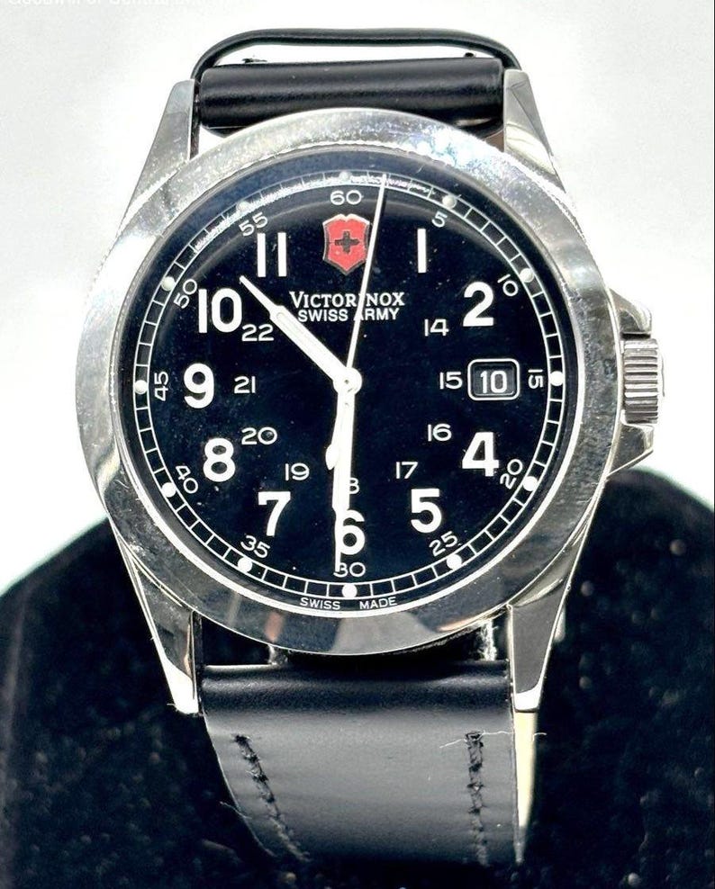 May include: A black-faced Victorinox Swiss Army watch with a stainless steel case and a black leather strap. The watch features white numerals, a date display, and the Victorinox logo. The watch is a classic design.